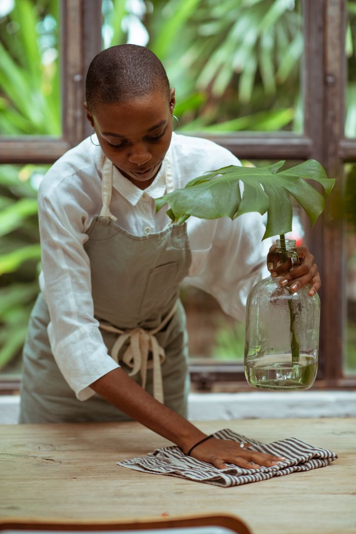 Concentrated African American waitress using microfiber wiping cloth to clean wooden table under stylish glass vase with green leaf while standing against big window and park