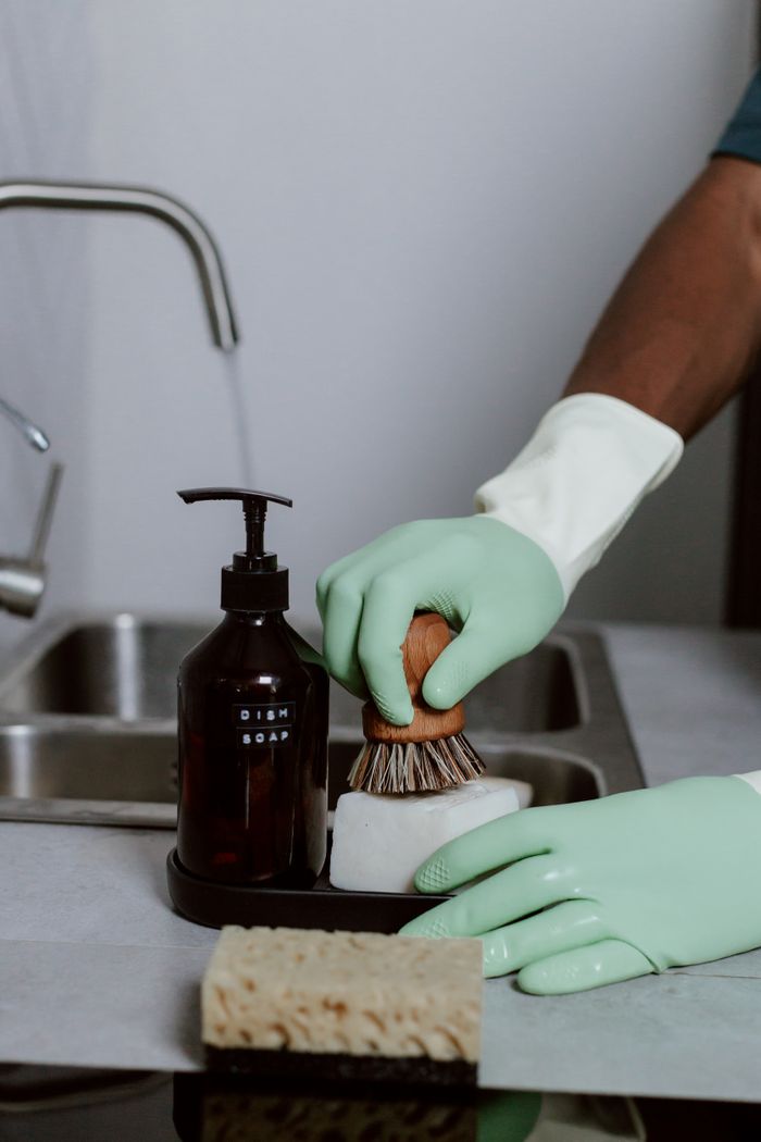 Close-up of hands in green gloves washing dishes with dish soap and brush in a kitchen sink.